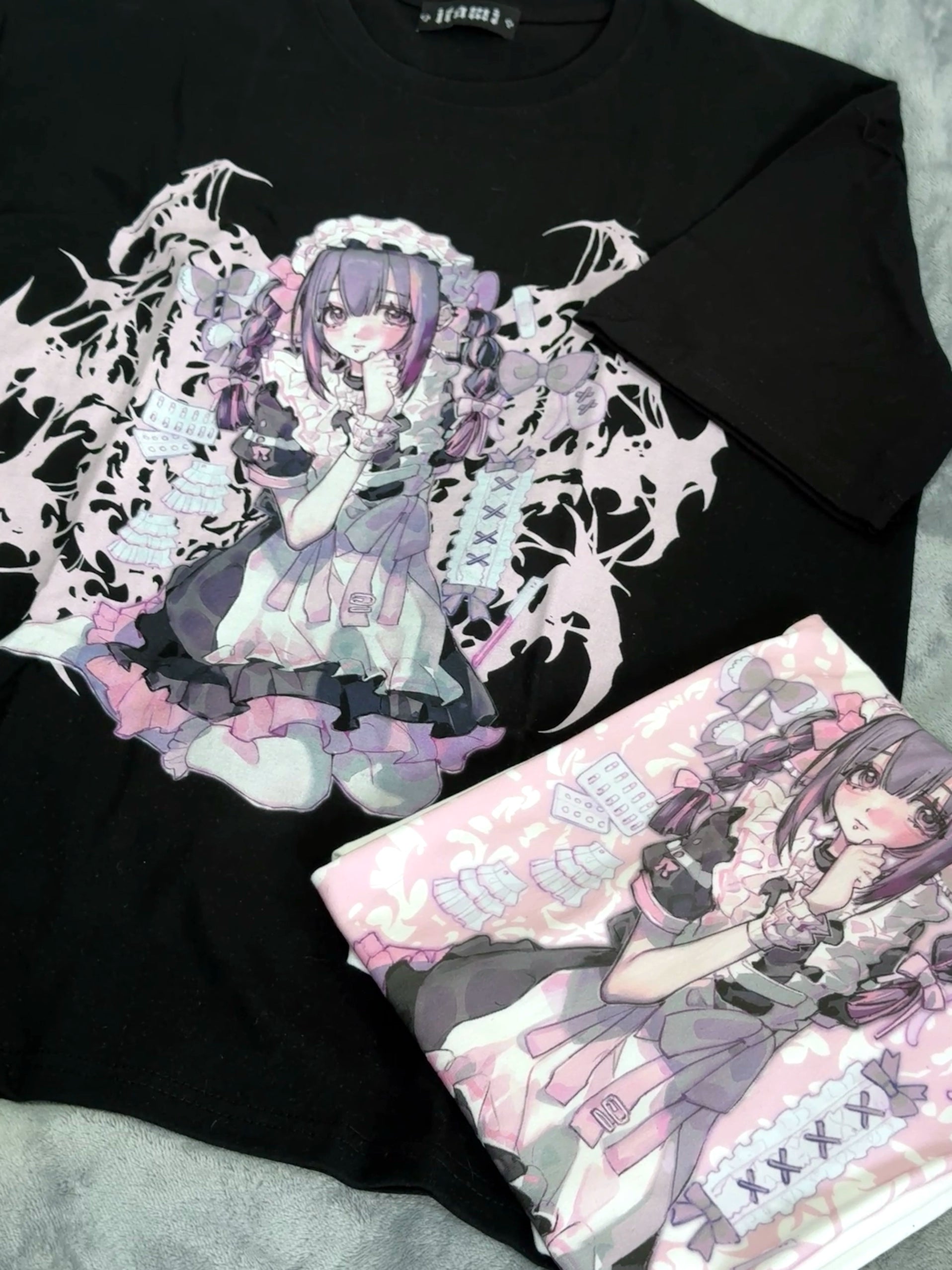 Flat lay overhead shot of folded black oversized Jirai Kei anime graphic t-shirt paired with folded pink variant on grey plush fabric background