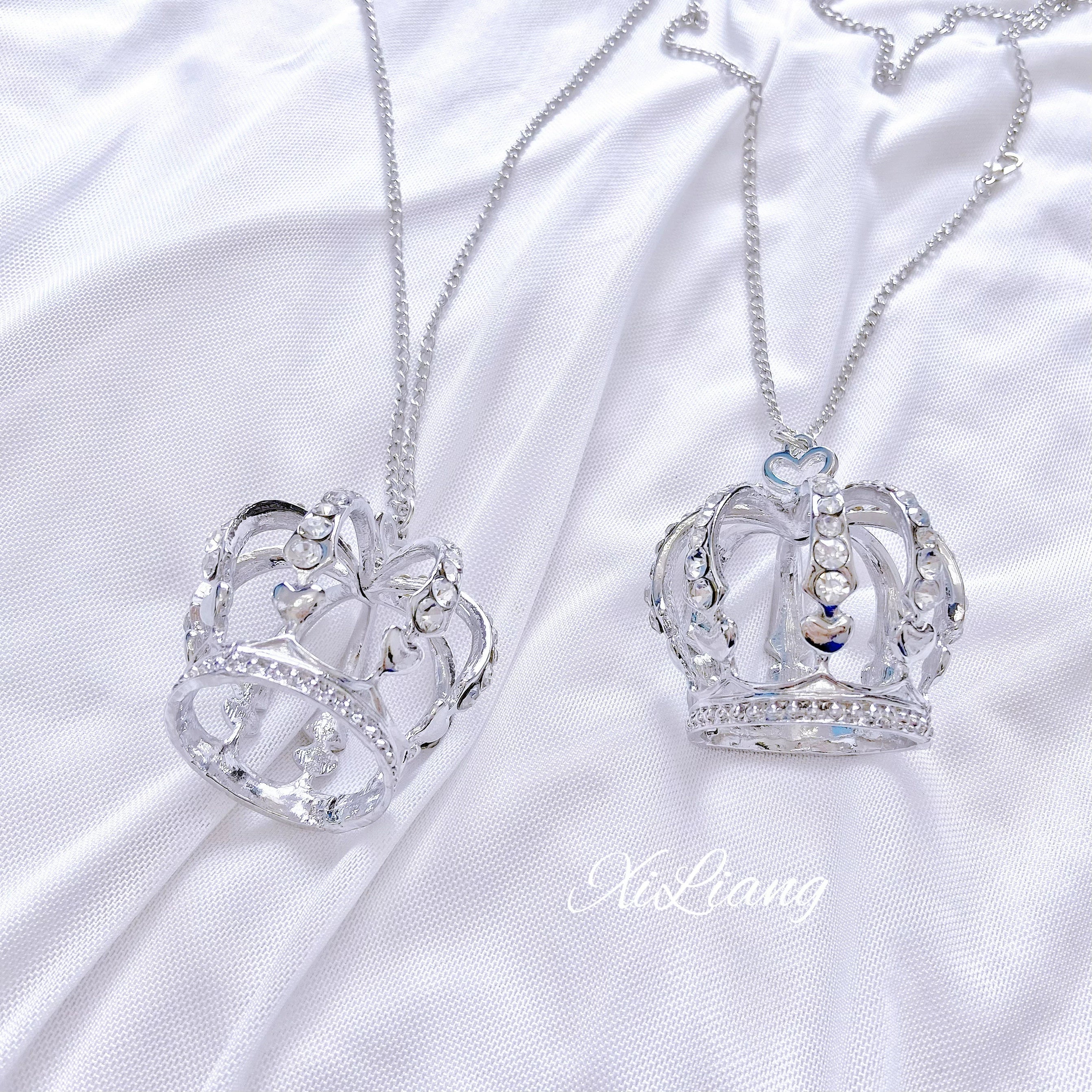 Pair of crown rhinestone necklaces on white satin, silver hollow crown designs with inlaid rhinestones, one with prominent top crystal details, matching set