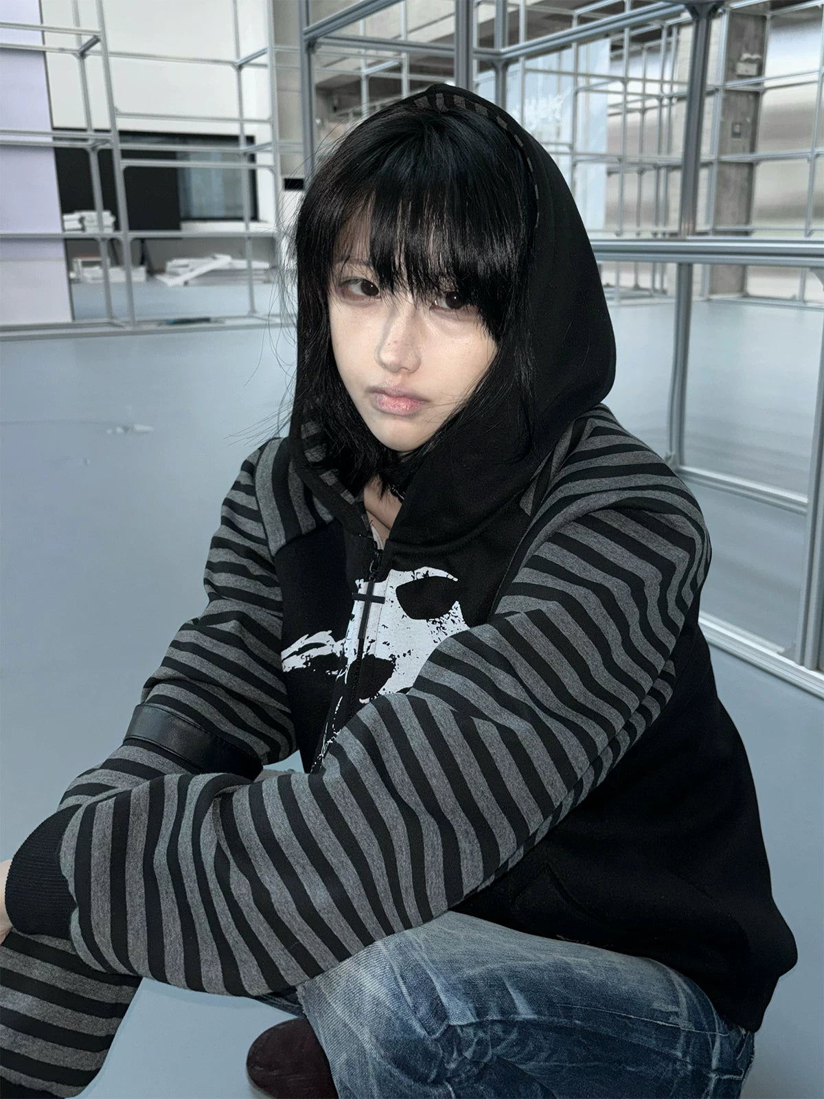 Female model in industrial indoor space wearing hooded scythe print zip coat with hood up grey striped sleeves and jeans side view profile