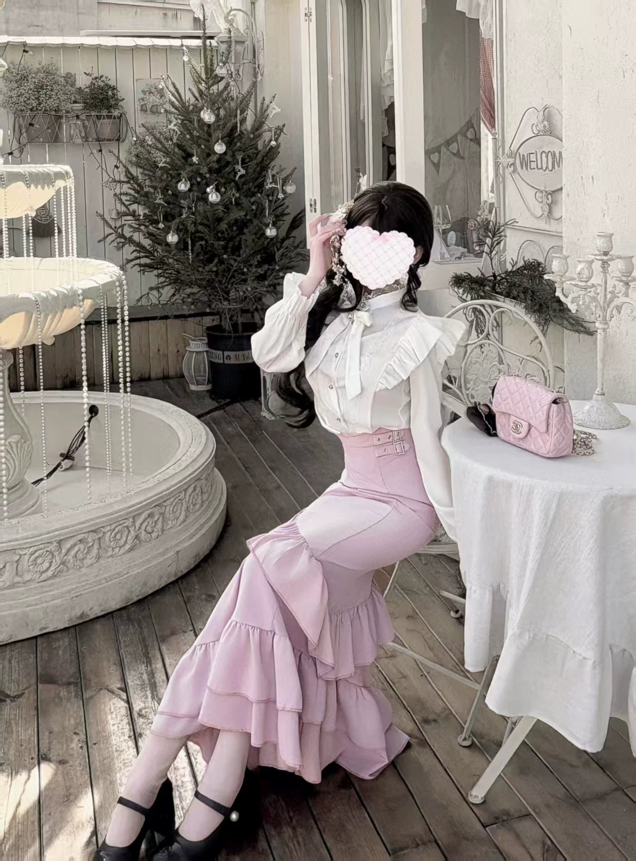 Model seated outdoors wearing romantic pink fishtail skirt and white blouse set with crystalline collar bow and pink crossbody handbag demonstrating Jirai Kei styling