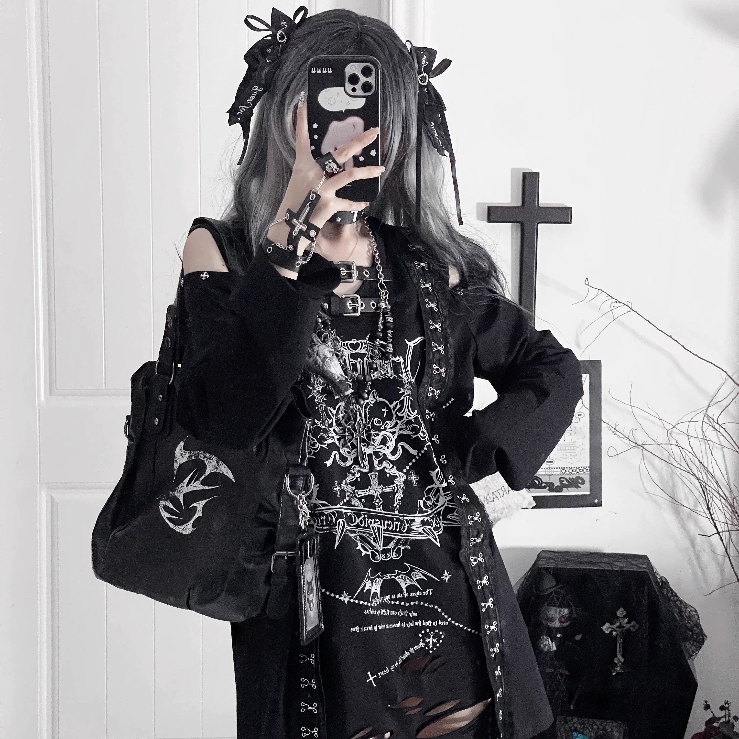 Mirror selfie full-body view of alternative girl wearing black goth long-sleeve tee with gothic prints and cut-out shoulders, accessorized with crossbody bag and hair bow for Jirai Kei aesthetic
