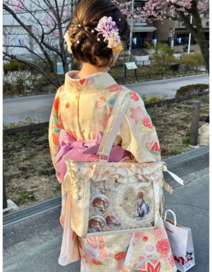 Beige satin Lolita itabag back view showing ruffled trim and decorative anime pins in clear window, paired with traditional kimono outfit on Japanese street