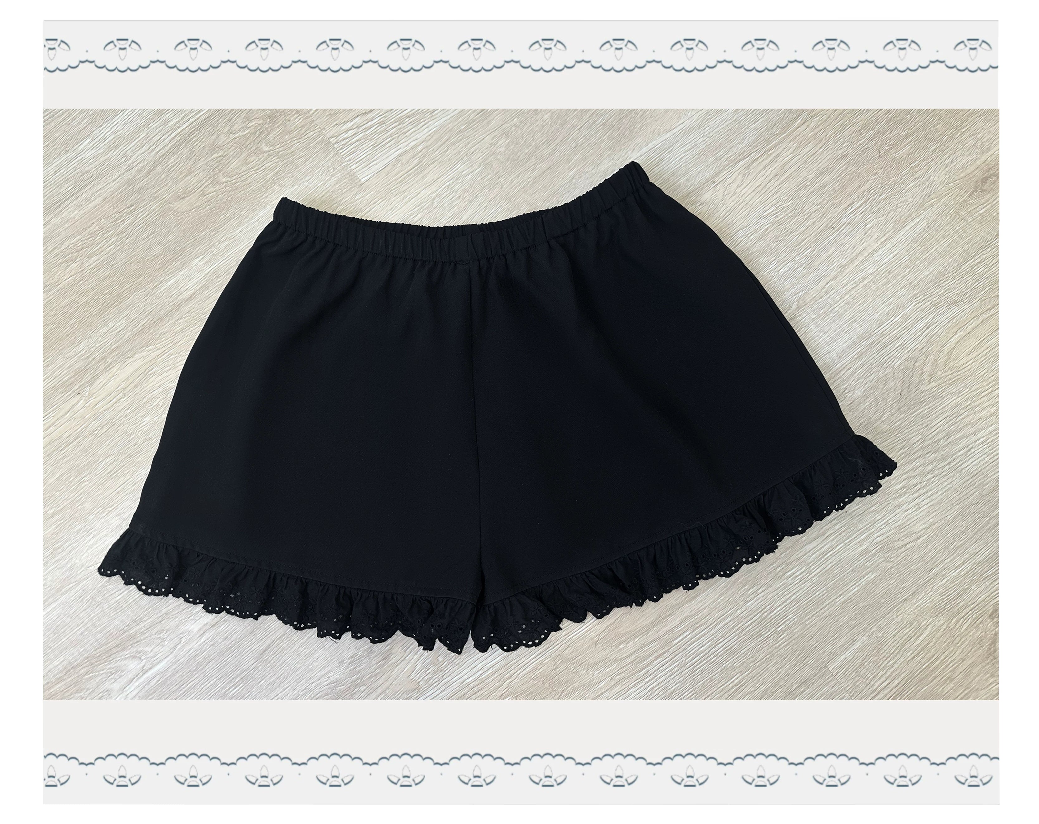Flat lay front view of black ruffled safety shorts with lace-trimmed hem and elastic waistband on wooden floor background