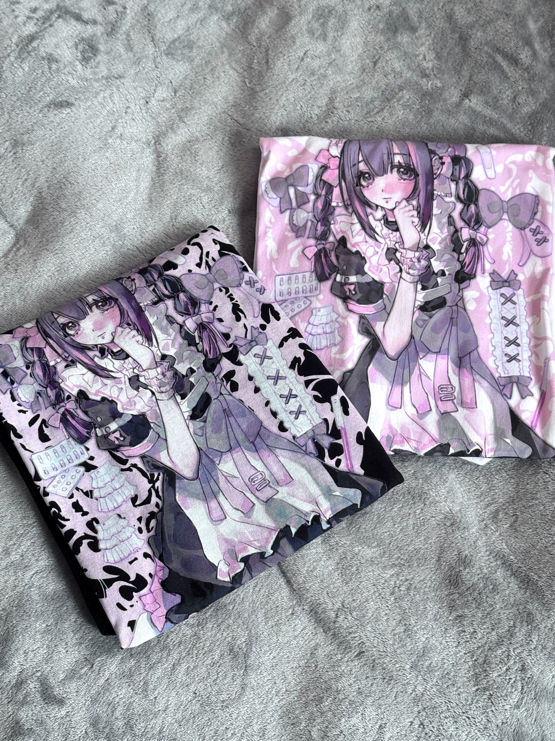 Overhead flat lay composition of two folded oversized Jirai Kei anime maid graphic t-shirts in different color bases showing matching print designs on plush grey background