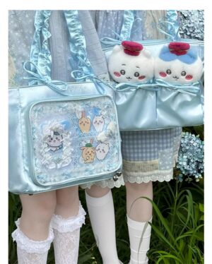 Light blue imitation silk Jirai Kei itabag with transparent badge display window and decorative bowknot straps worn by model in sweet Lolita attire outdoor garden setting