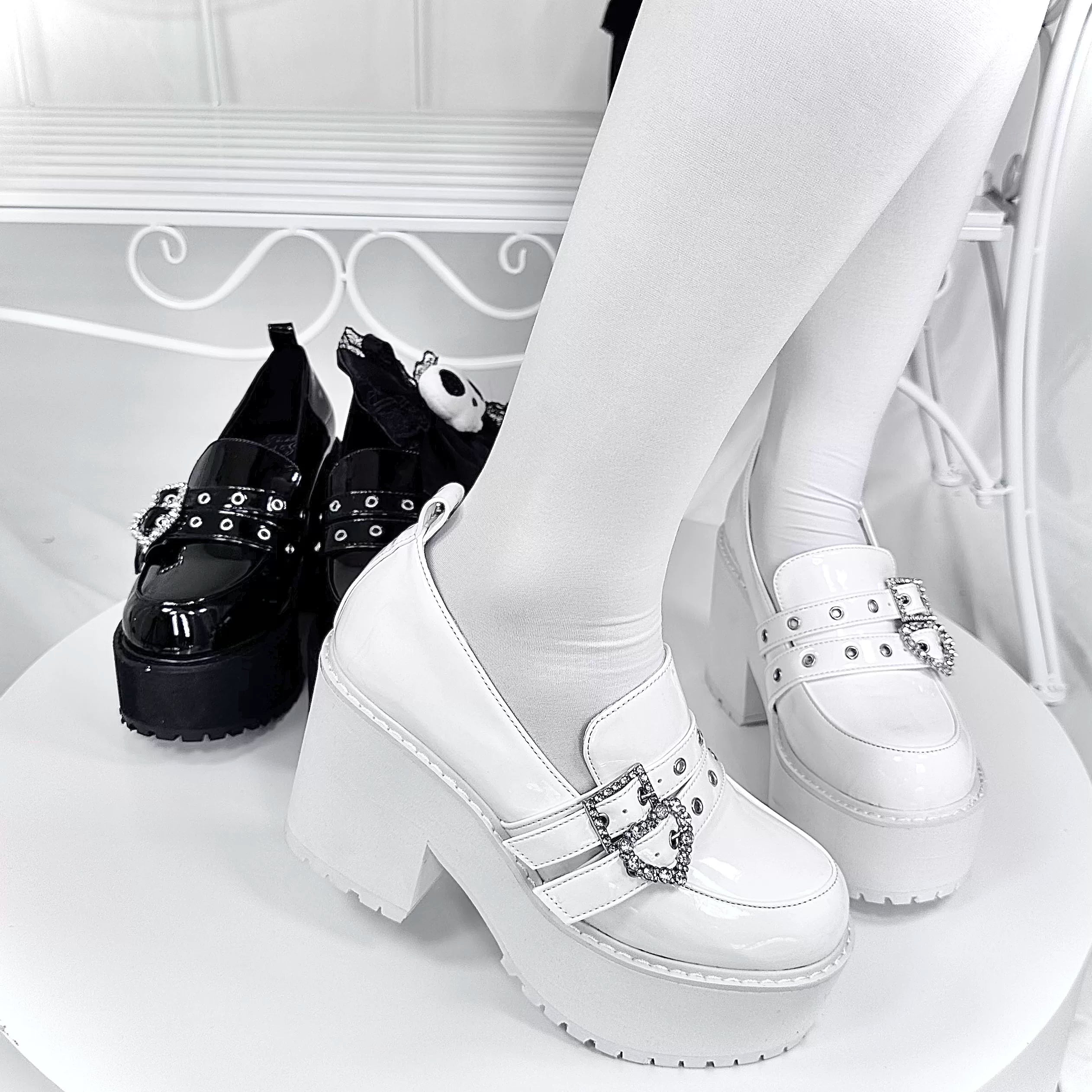 Dual-tone patent leather platform loafers three-quarter overhead angle on model in white stockings with heart rhinestone buckles and wrought iron furniture backdrop