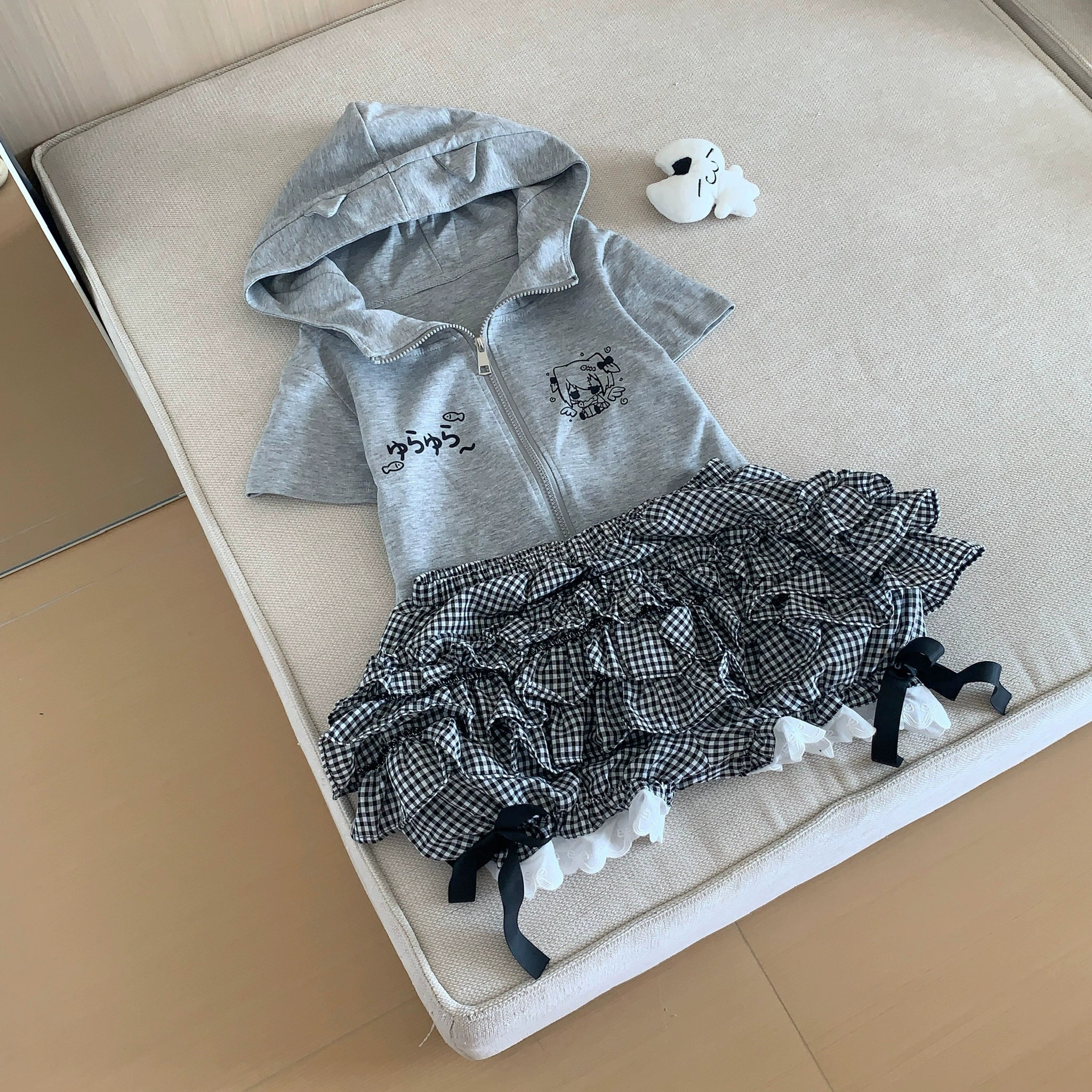 Flat lay product display of complete Jirai Kei outfit on soft cushion including hooded grey zip-up sweatshirt with cartoon graphics matching black white ruffled plaid mini skirt built-in lace shorts loafers and accessories