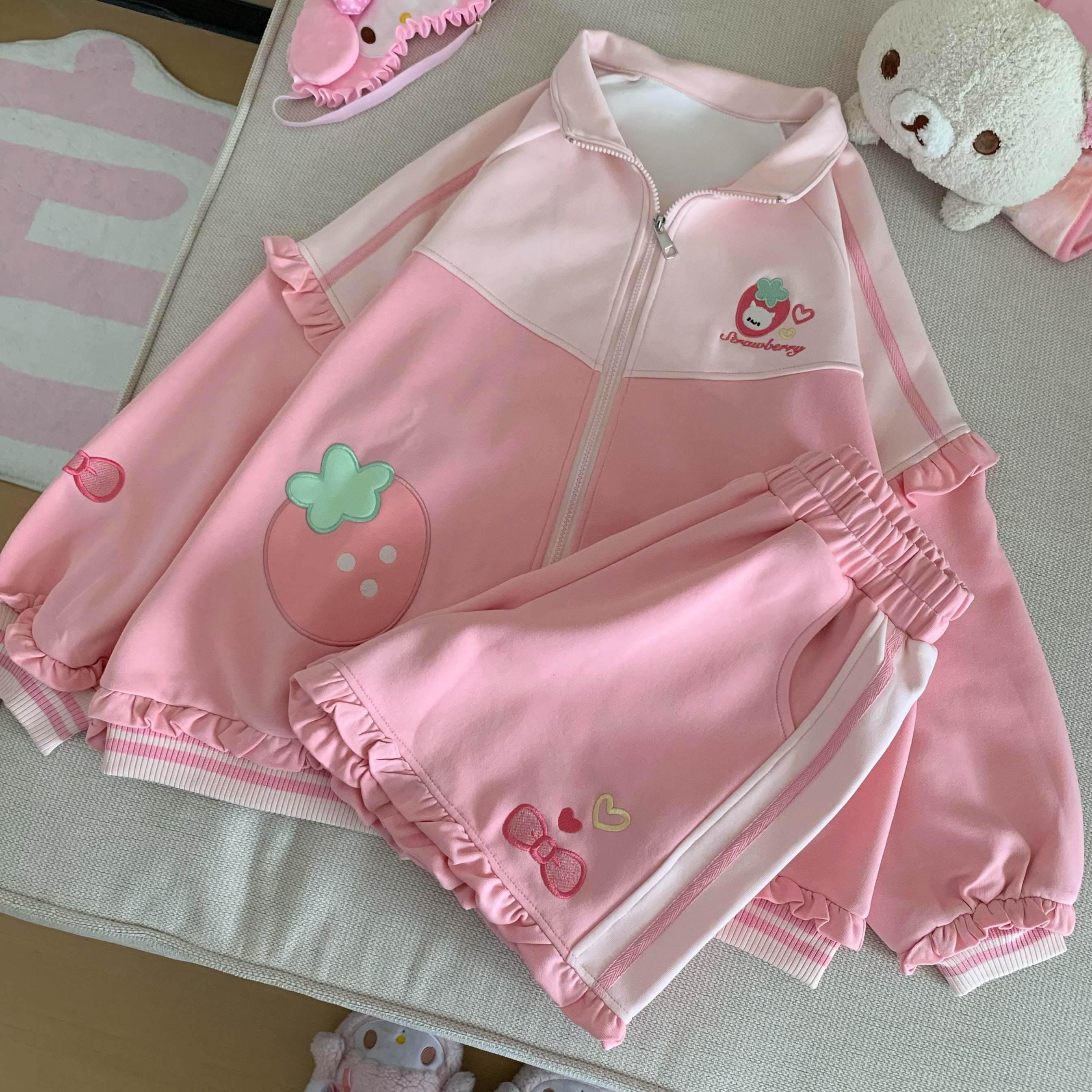 Bedroom styled flat lay of pink Jirai Kei strawberry two-piece set with elastic waist shorts and plush toy accessories, creating kawaii aesthetic lifestyle content