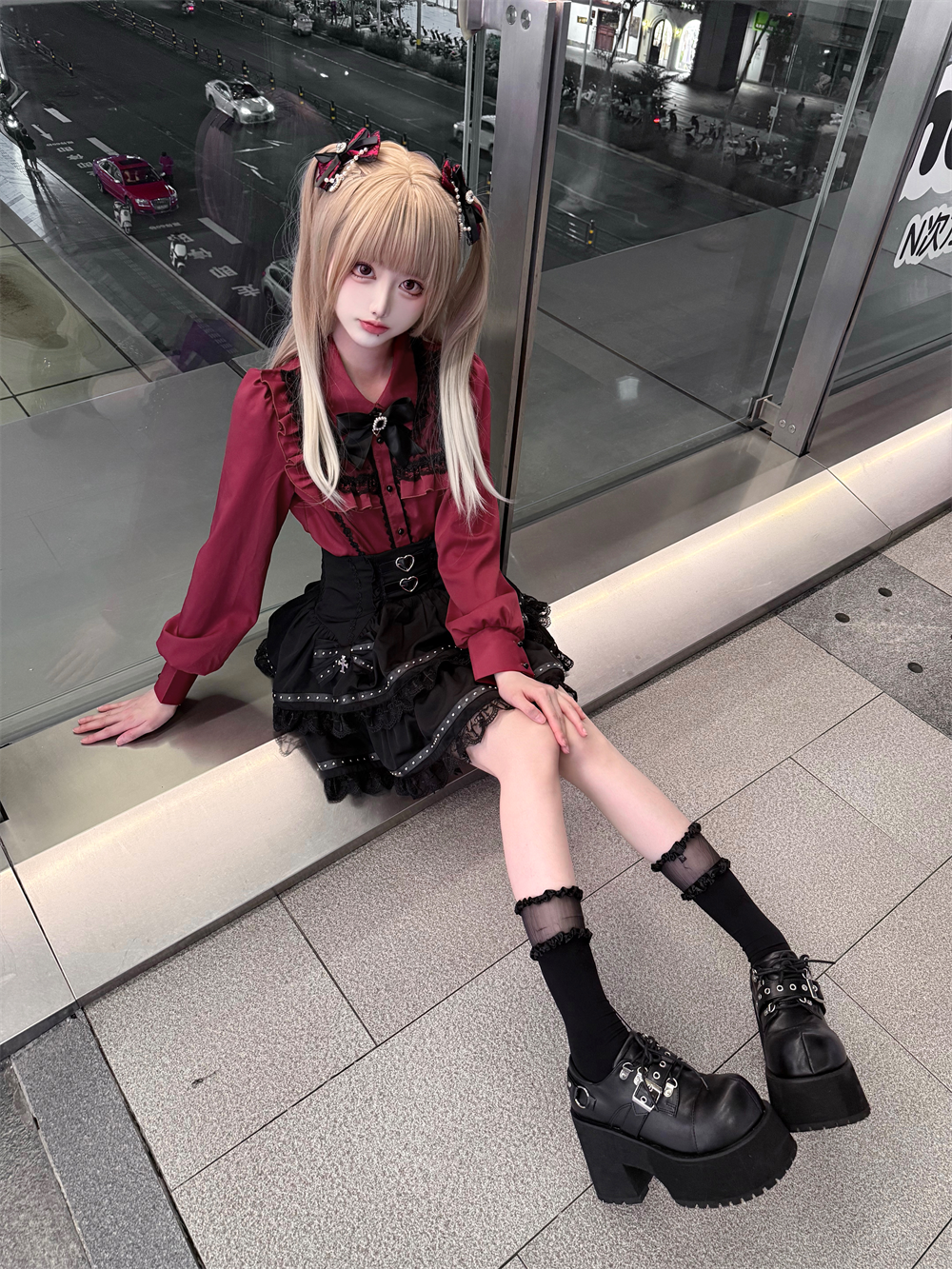 Seated side view of Jirai Kei fashion showing relaxed sitting pose wearing matching wine-red blouse and black tiered lace skirt on nighttime commercial terrace