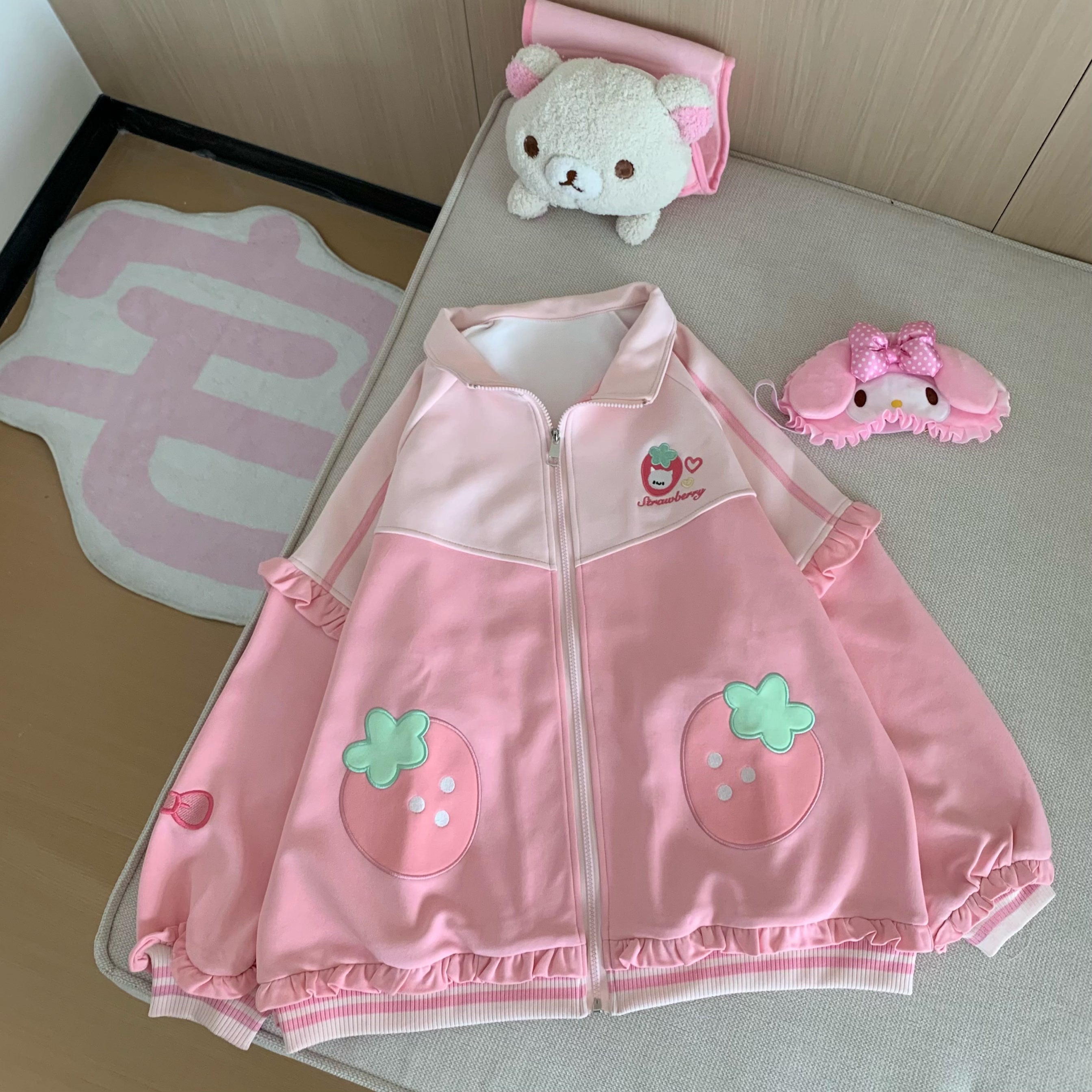 Top-down flat lay of pink strawberry Jirai Kei coat featuring stand collar, ruffled sleeve edges, and detailed strawberry embroidery patches, styled with cute hair accessory