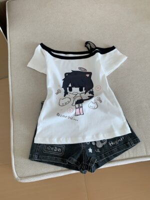 Off-shoulder Jirai Kei anime catgirl t-shirt with black cold-shoulder trim and white base, paired with dark denim shorts featuring text graphics, flat lay on upholstered cushion