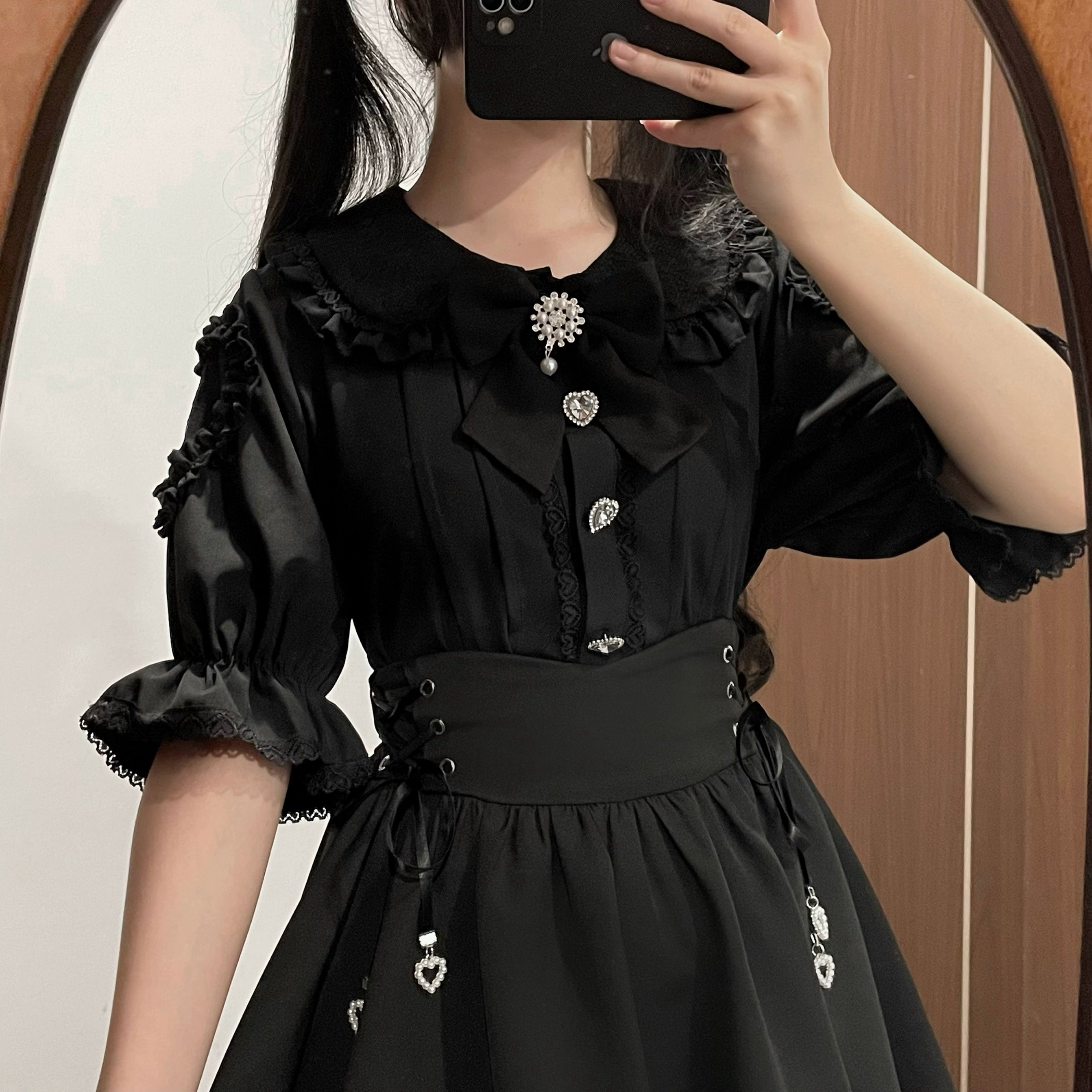 Black Jirai Kei blouse and skirt set mirror selfie close-up on girl model, highlighting ruffled puffy sleeves, heart-shaped buttons, pearl bow tie, crystal brooch, and lace detailing - fashion detail showcase