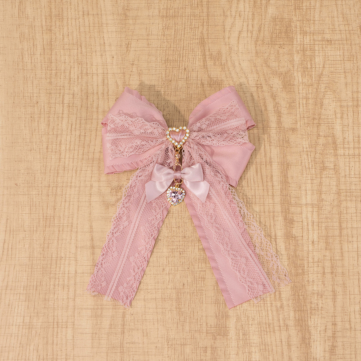 Dusty pink Jirai Kei lace double bow brooch with sparkling rhinestone heart charm, handcrafted satin ribbon detail, flat lay on wooden surface