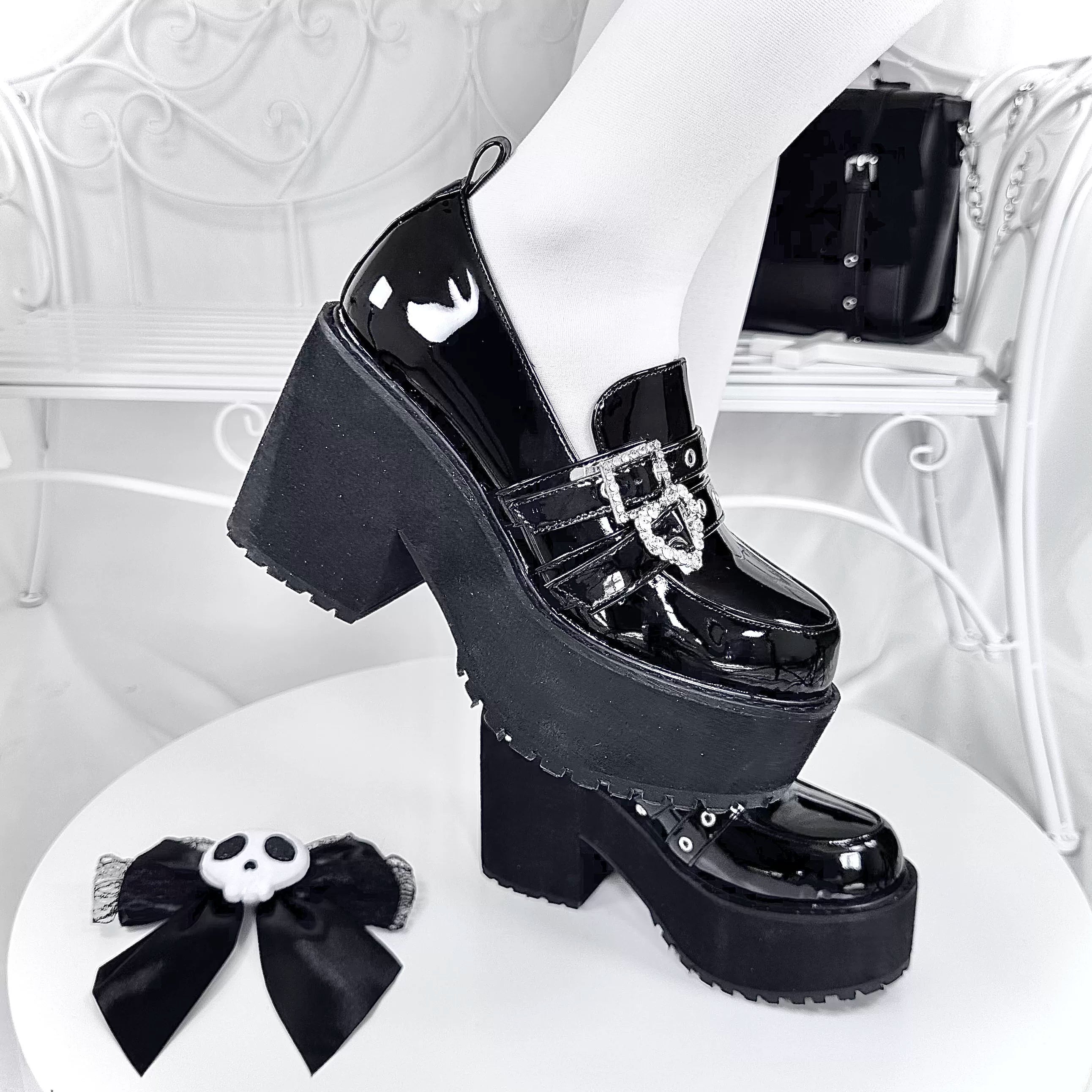 Side profile close-up of black patent leather platform loafers showing chunky platform sole heart rhinestone buckle and skull bow decor on vintage white furniture