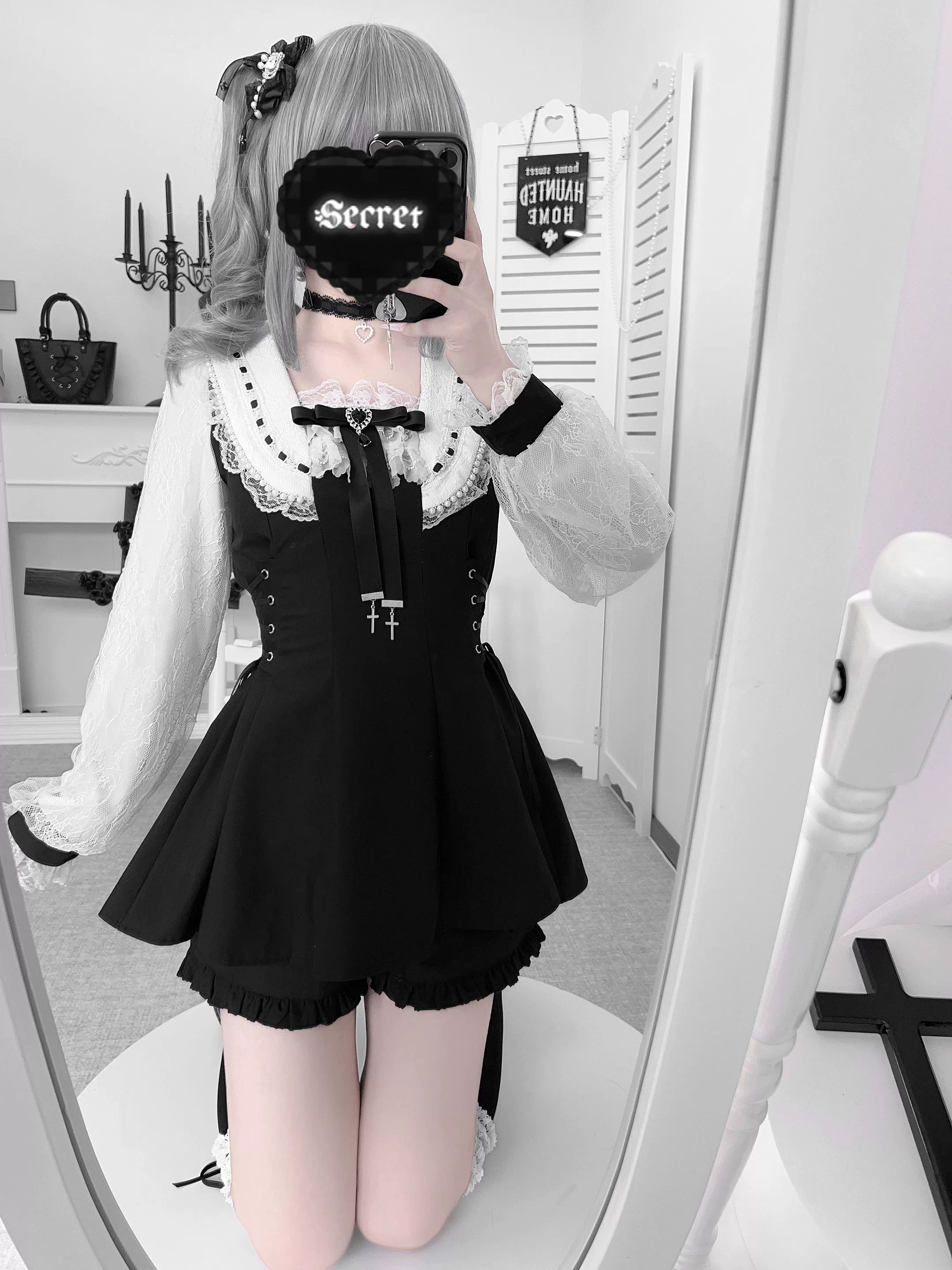 Kneeling mirror selfie of model in solid black Jirai Kei outfit showing ruffled blouse cuffs and heart-themed phone cover detail
