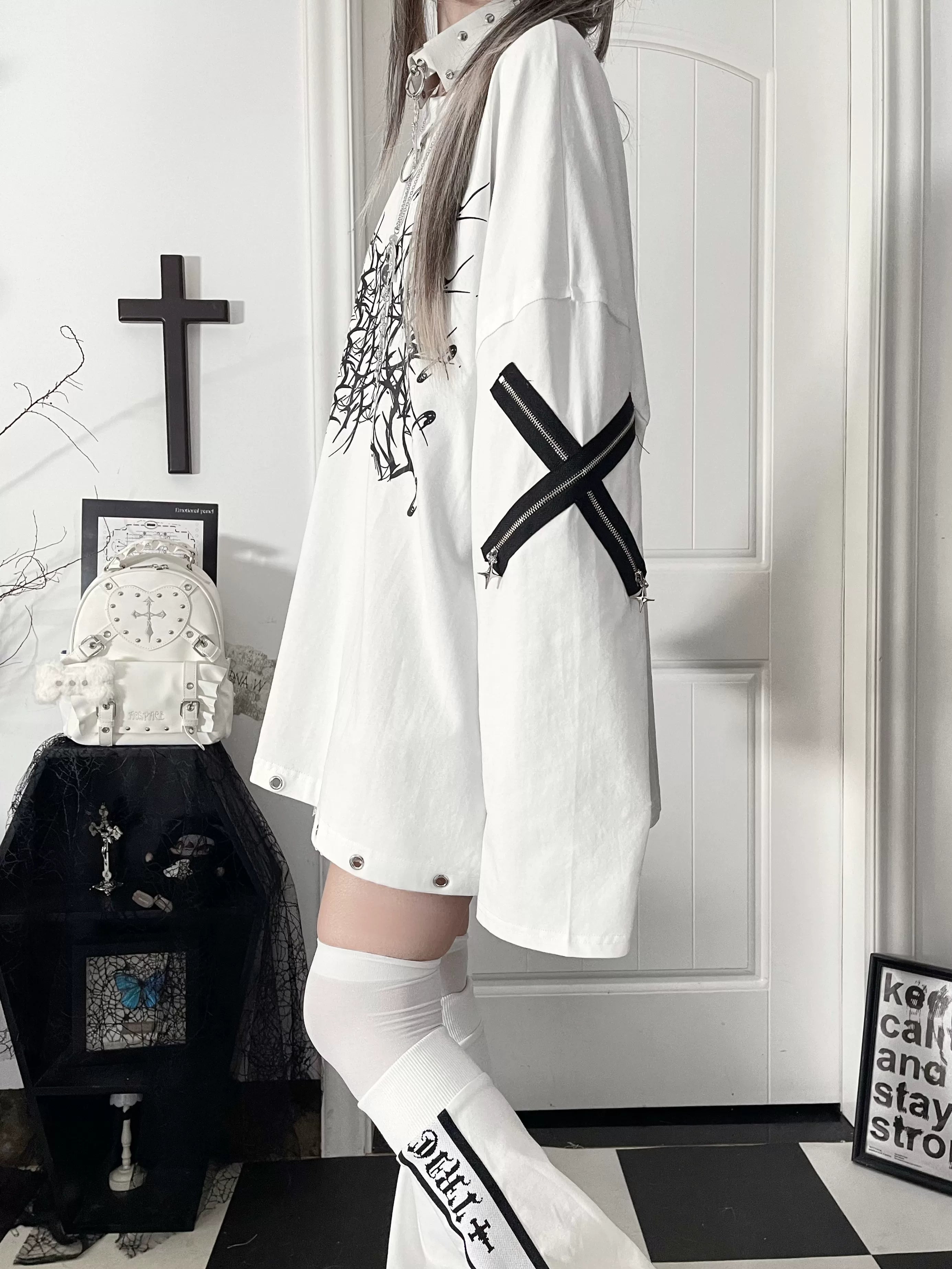 Side profile view of white jirai kei oversized gothic long-sleeve tee featuring black thorn cross print with cross strap sleeve construction, worn in gothic indoor environment