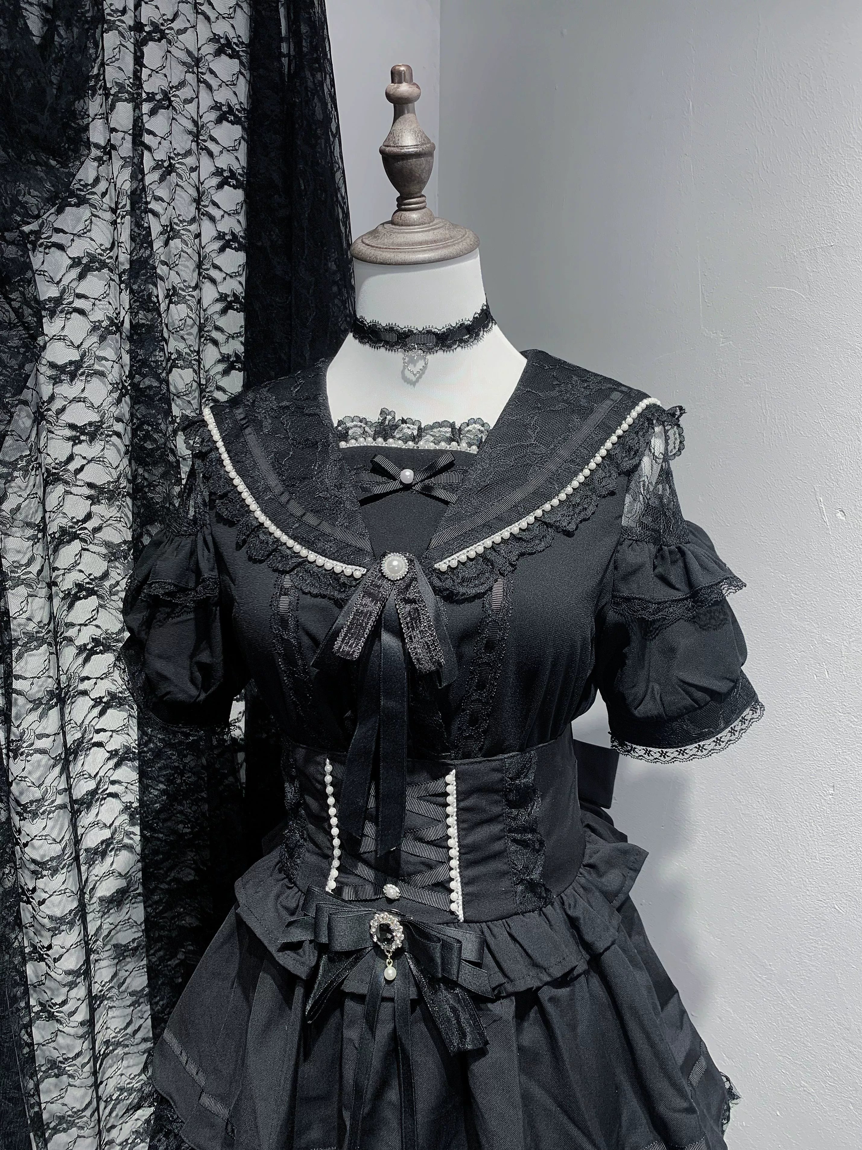 Short-sleeved black Jirai Kei sailor collar blouse featuring structured neckline with lace detailing, removable bow accent, pearl button fasteners, and gothic-inspired design elements in studio view