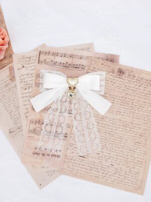 White lace bow heart rhinestone hair clip with long flowing lace tassels and sparkling heart pendant displayed on vintage letters and sheet music background, showcasing Jirai Kei double ponytail accessory design from overhead flat lay perspective