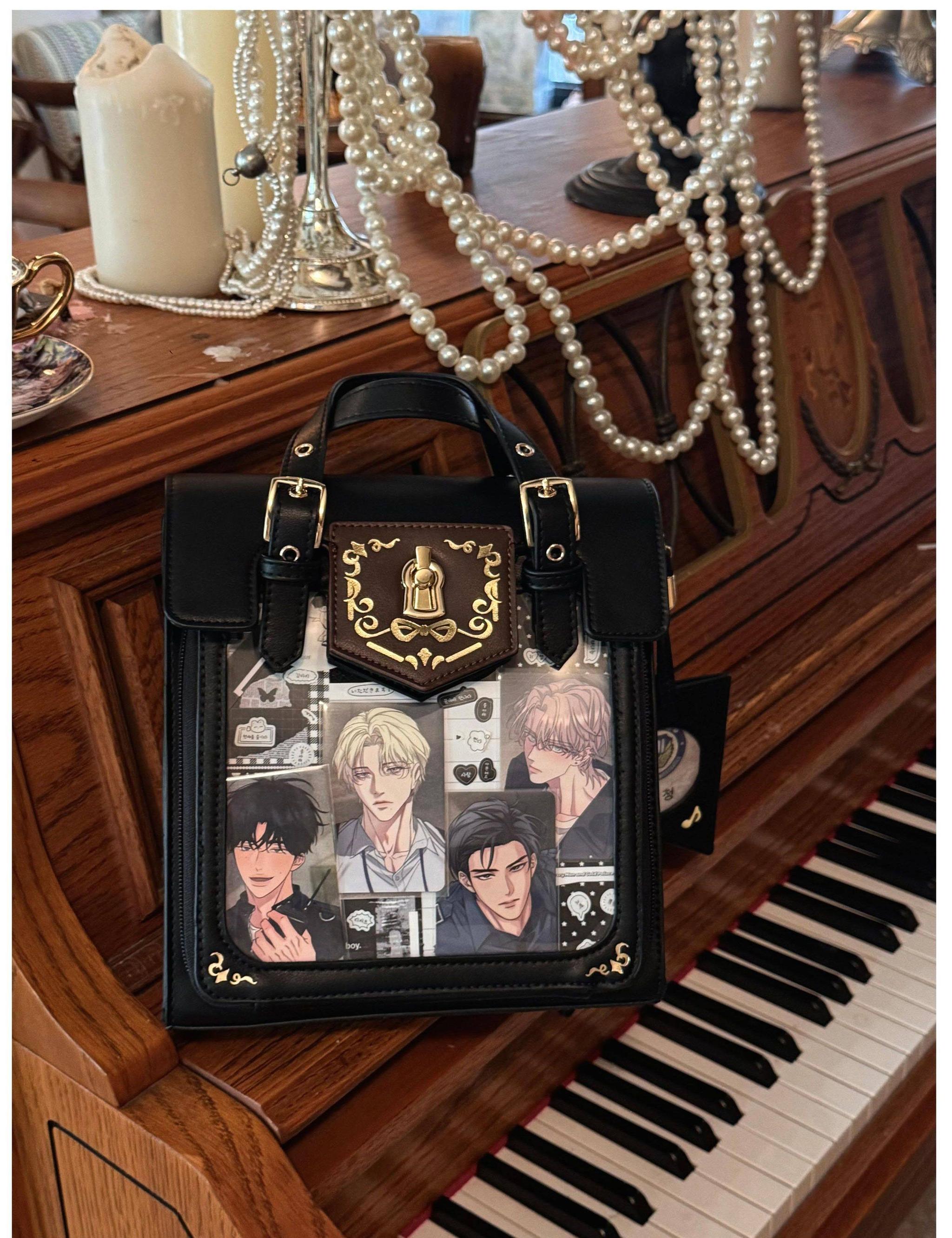Black vintage Jirai Kei ita satchel bag with ornate gold royal crest hardware and clear window displaying multiple anime character portraits, indoor vintage piano table aesthetic