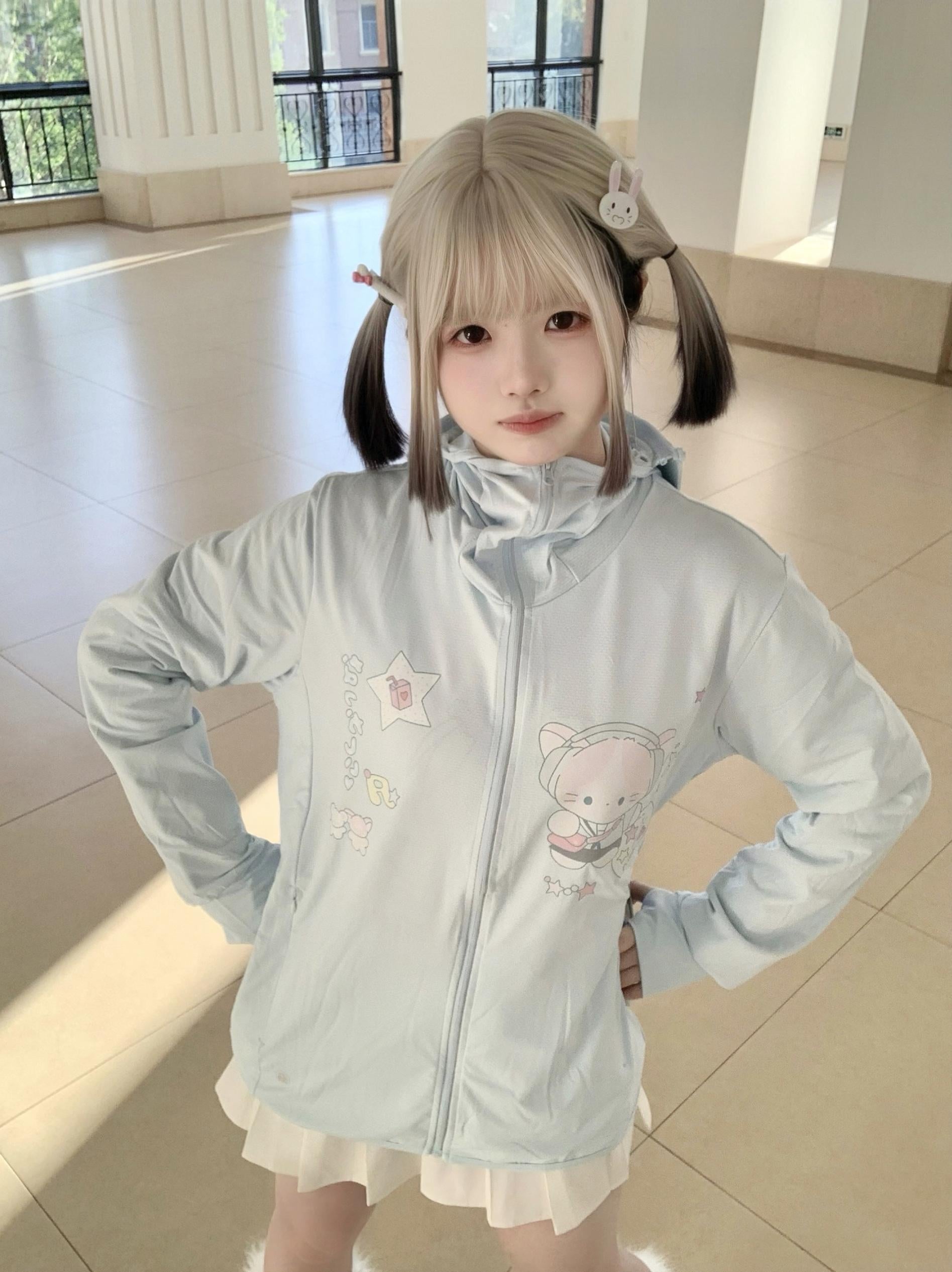 Jirai kei UV protection jacket with detachable hood on blonde model with hands on hips, hooded zip-up style with cartoon prints visible, bright window backdrop