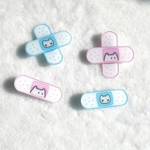 Four-piece bandage shaped hair clips set with cat detail in pink and water blue, flat lay on white fluffy fabric showing cross and straight stripe styles for Tenshi Kaiwai and Jirai Kei fashion