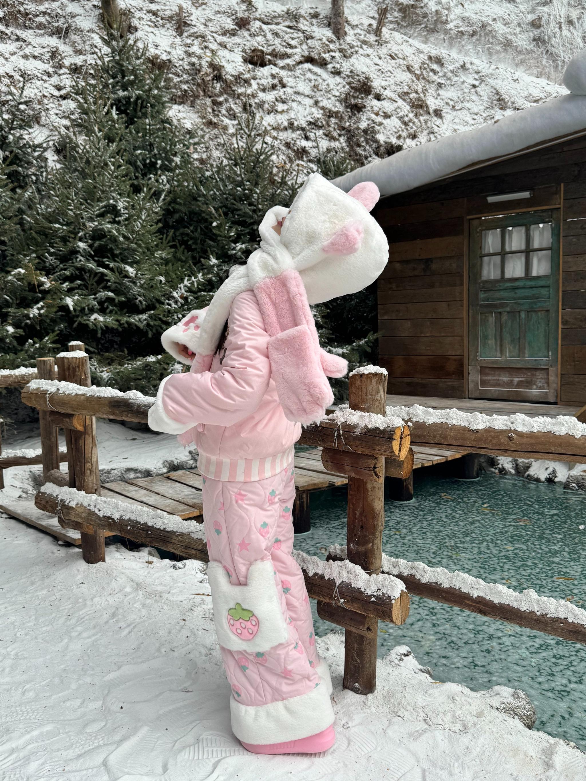 Back side view of pink Jirai Kei padded coat with adjustable bear ear hood, fuzzy trim, pink platform shoes, strawberry patch quilted pants at snowy wooden bridge