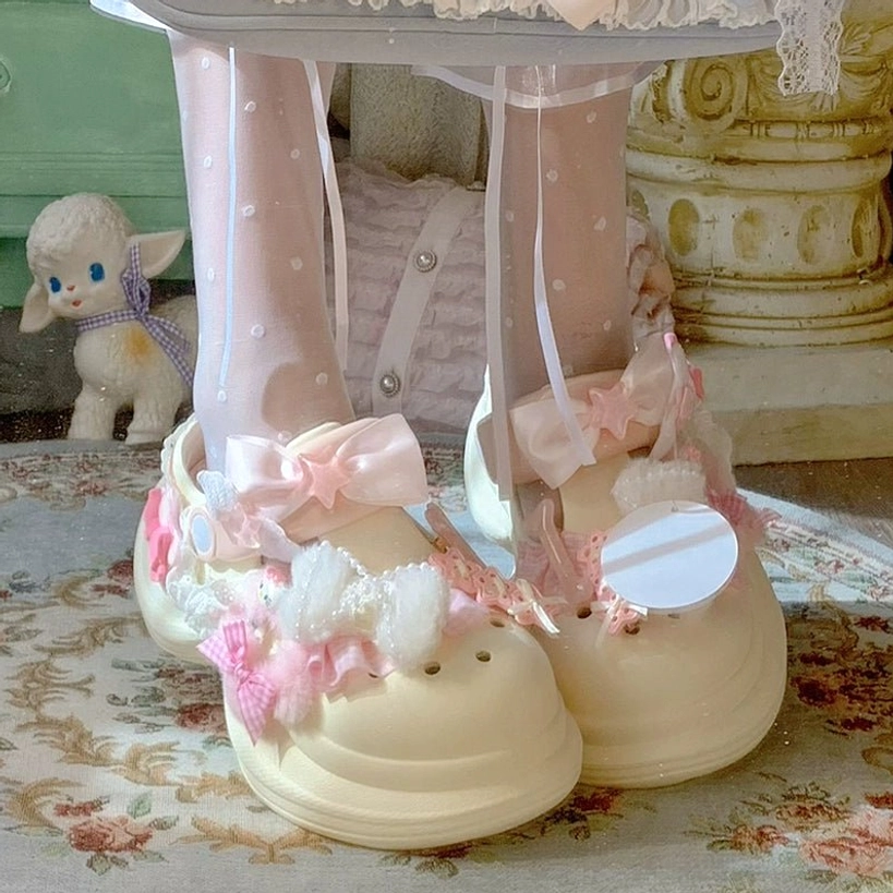 Beige thick-soled kawaii clog sandal with pink bows, fuzzy pom-poms, round badge charm, frontal full foot view with sheer polka-dot ribbon socks