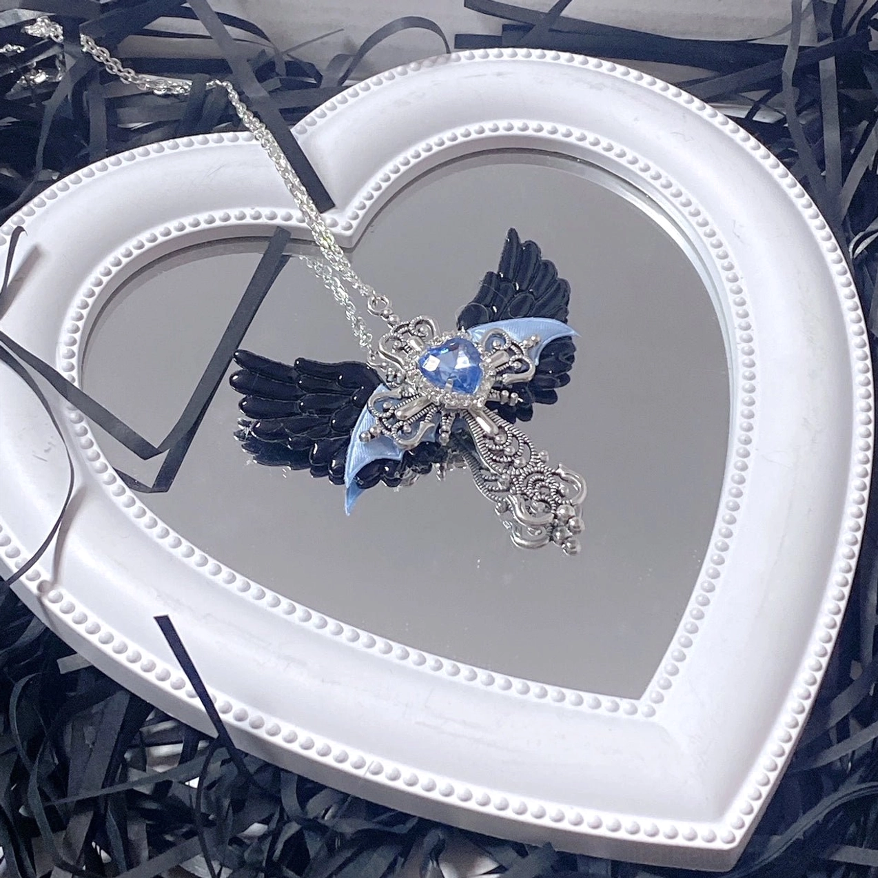 Black and blue Y2K Jirai Kei winged cross necklace featuring blue heart gem and silver cross on white heart-shaped mirror frame