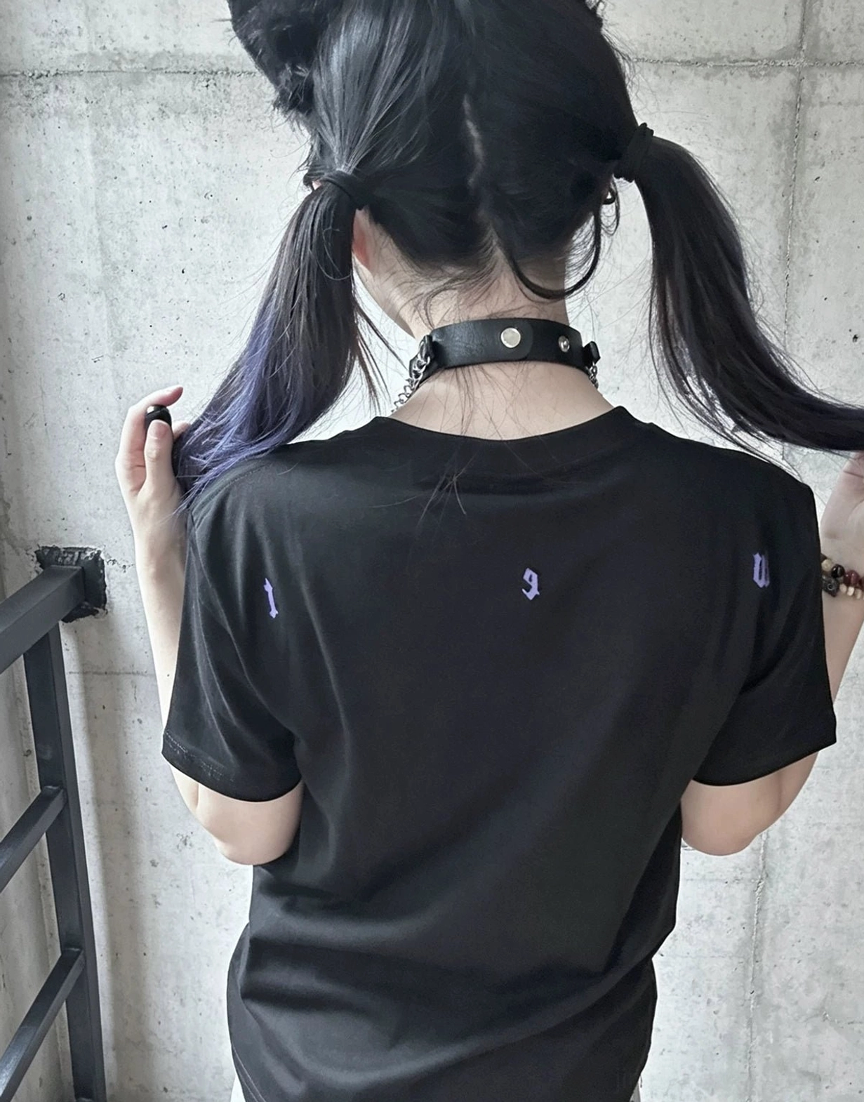 Back view of black cross print t-shirt, female model with twin tails hairstyle, tiny purple decorative prints visible on back hem, concrete wall background
