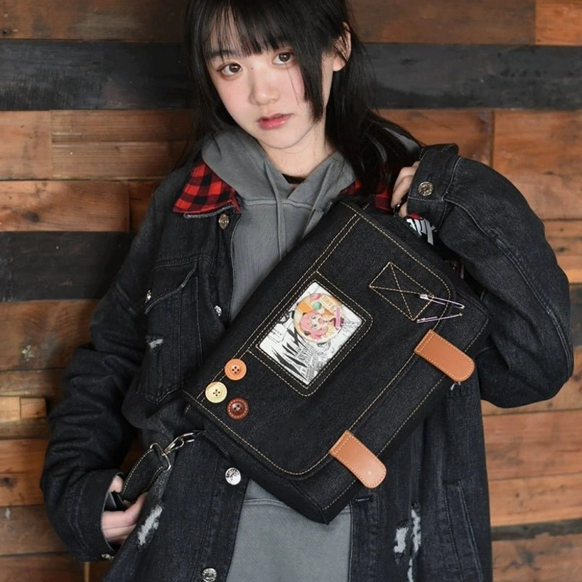 Black denim itabag crossbody bag side front angle with model in denim jacket and gray hoodie, anime print display window with decorative buttons and brown leather fasteners