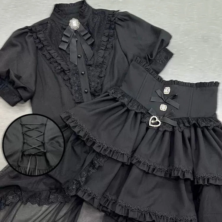 Black gothic shirt and high-waisted skirt flat lay detail view featuring puff sleeves, lace ruffle front, corset lace-up back, layered ruffle skirt with heart buckle