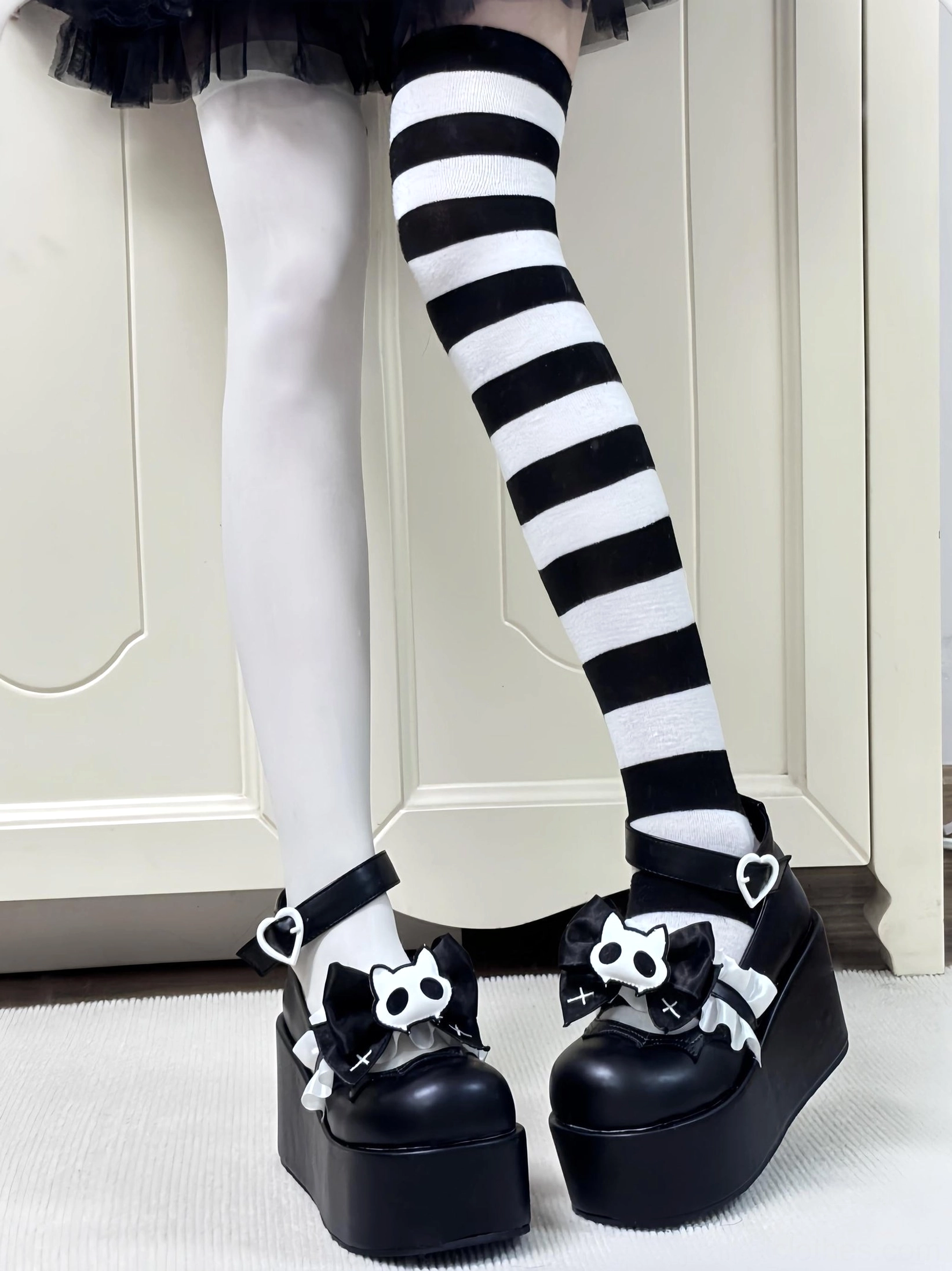 Full front leg view of black gothic platform shoes with silver heart buckle and mismatched stockings indoor setting
