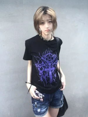 Black gothic t-shirt with purple thorn cross graphic print on front, female model wearing with denim shorts against concrete wall, dark aesthetic subculture style