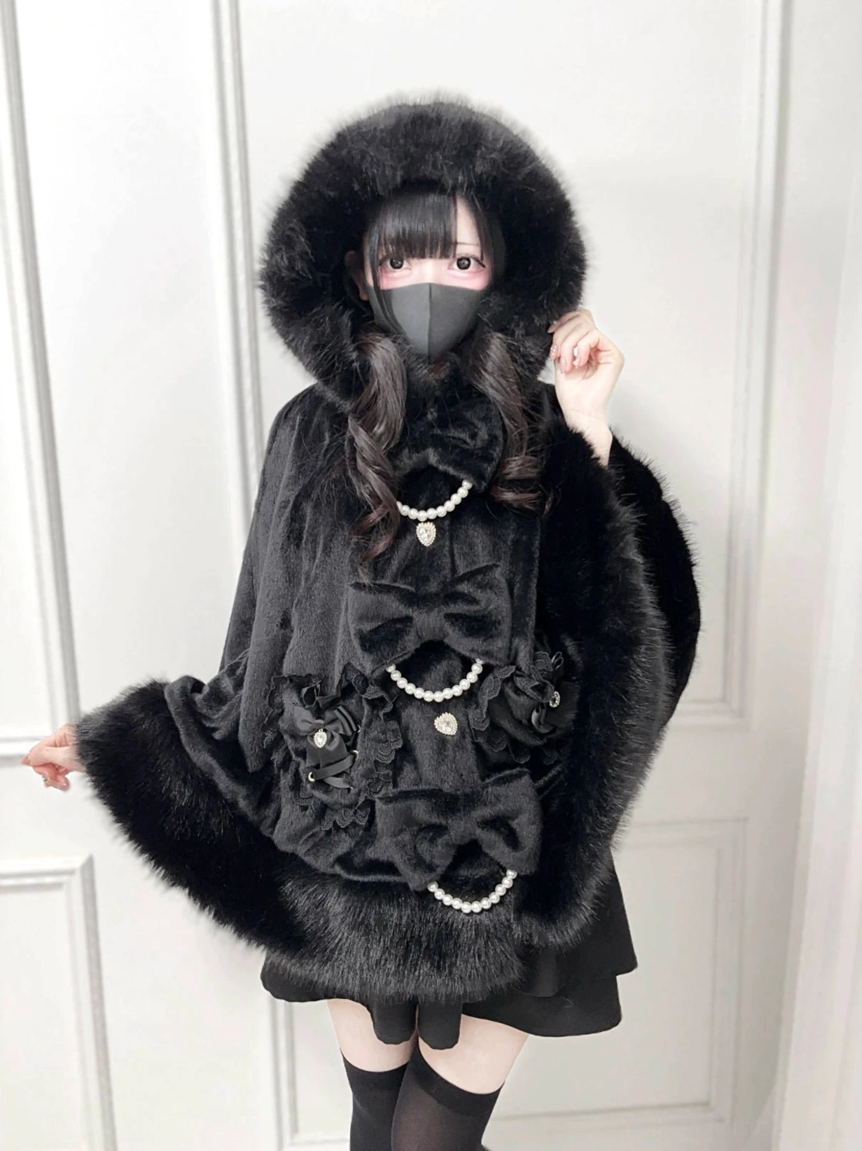 Black hooded Jirai Kei winter cape with faux fur trim and pearl bow decorations, front three-quarter view on female model