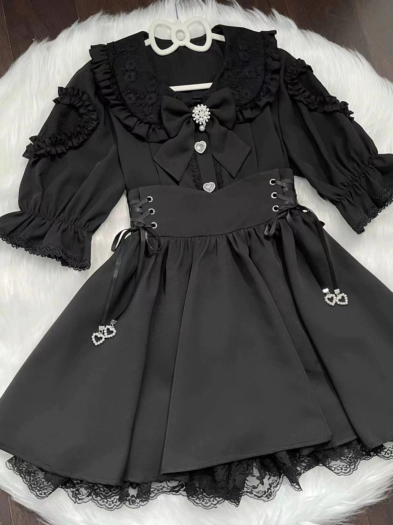 Black Jirai Kei blouse and skirt set flat lay front view on white plush background, featuring delicate lace trims, corset side lacing details, heart pendant charms, and crystal brooch accessories - high fashion underground Japanese streetwear
