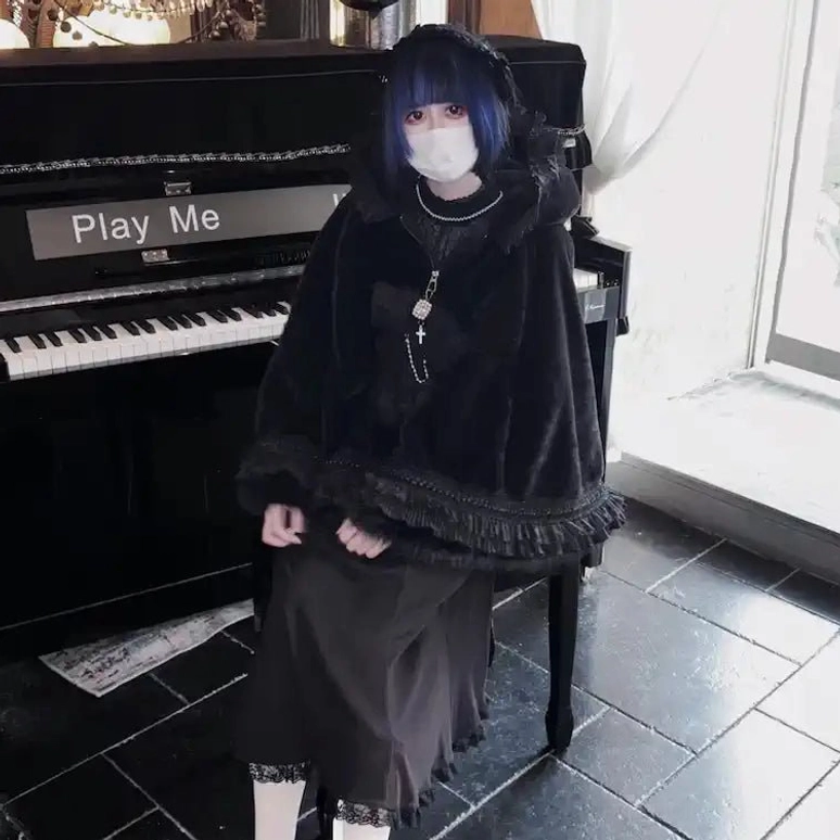 Direct front view of black cat ear fleece cape hoodie with fuzzy plush hem, long pendant necklace, ankle-length black skirt at piano cafe setting