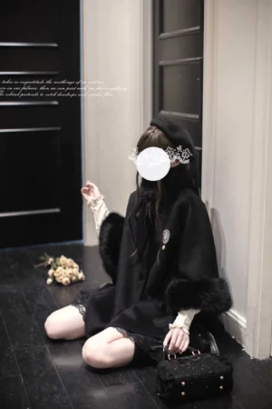 Overhead view of black wool-blend jirai kei cloak showing faux fur cuffs and loose cape silhouette on seated model indoors