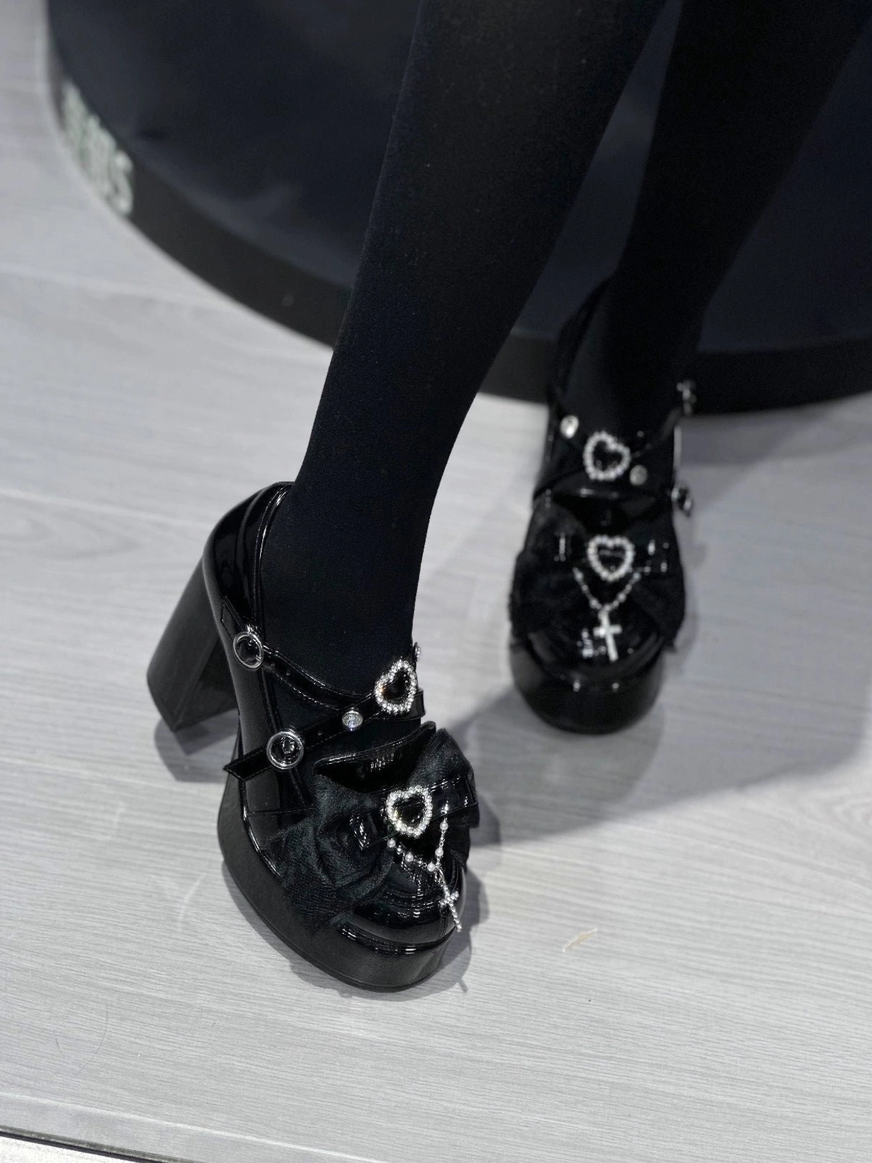 Side-rear angle of black patent Jirai Kei platform high-heel shoes on model in opaque tights, featuring heart rhinestone cross ornament, chunky block heel, and platform sole