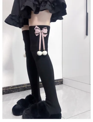 Black Jirai Kei over-the-knee plush socks with pink satin bow and white pom-pom charms, side front view on model wearing black ruffle skirt and fuzzy slippers, ribbed knit texture detail