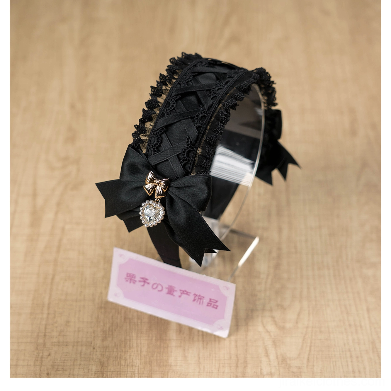 Black Jirai Kei lace headband close-up front three-quarter view on wooden table with product tag, highlighting large rhinestone pendant, frilled lace base, and satin bow construction details