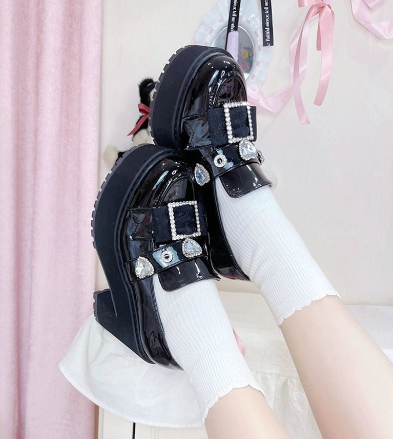 Black patent leather Jirai Kei platform loafers close-up side view with frilled white socks on pink fabric backdrop showing shiny finish and rhinestones