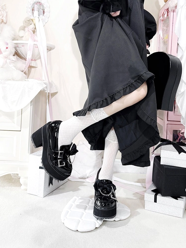 Black patent leather Jirai Kei platform shoes back side elevated angle with one foot lifted, showing square platform heel, rhinestone decor, glossy leather finish, side buckle, white socks, and black long ruffled skirt