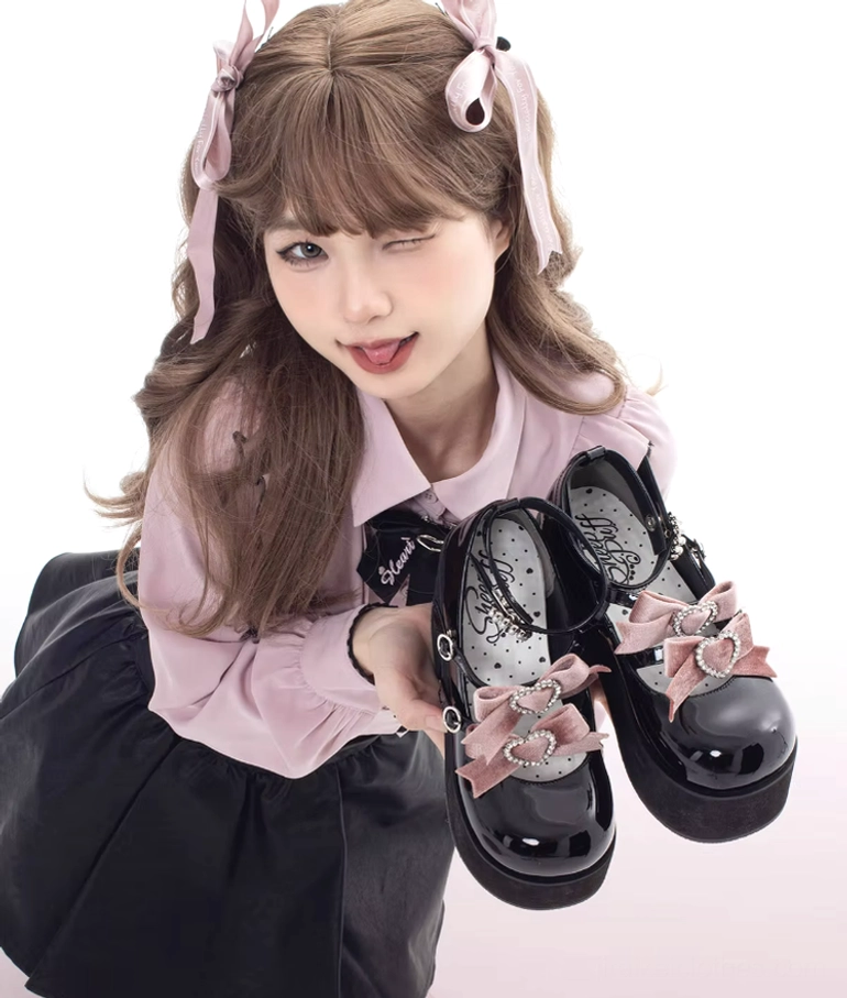 Sweet lolita model holding black chunky platform shoes with pink satin bow and rhinestone accents, wearing coordinating pink hair accessories and blouse