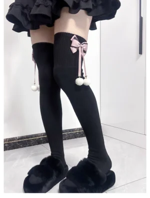Black over-the-knee Lolita plush socks with pink bow ornament and white pom-pom pendants, front view on model in black ruffle skirt, showing ribbed knit fabric and ankle decoration