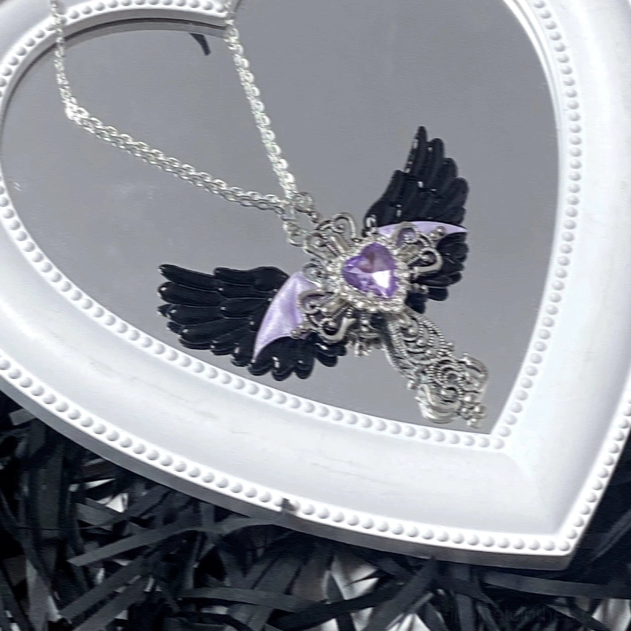 Black and purple Jirai Kei winged cross necklace with purple heart gem and silver cross on white heart mirror with dark shredded filler