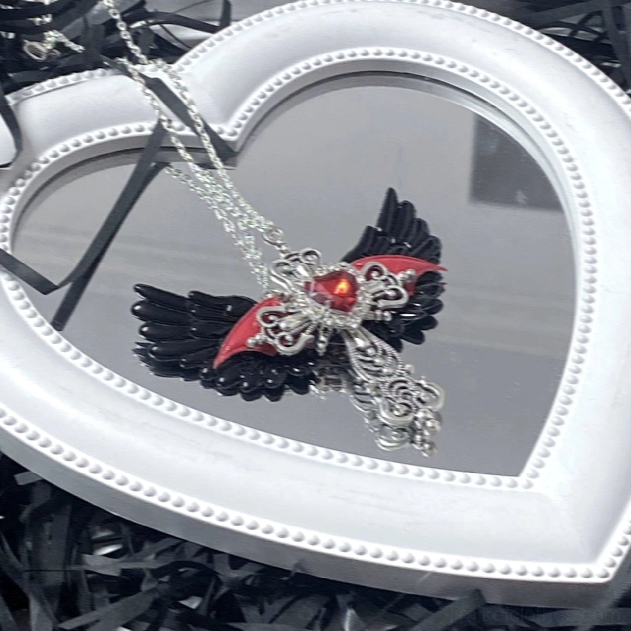 Black and red devil-style winged cross necklace with red heart rhinestone on white heart-shaped mirror frame against dark background