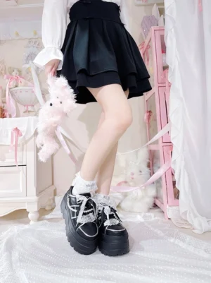 Black platform sneakers with rhinestone bow decorations worn by girl in black pleated skirt with white lace socks in pink bedroom Jirai Kei style