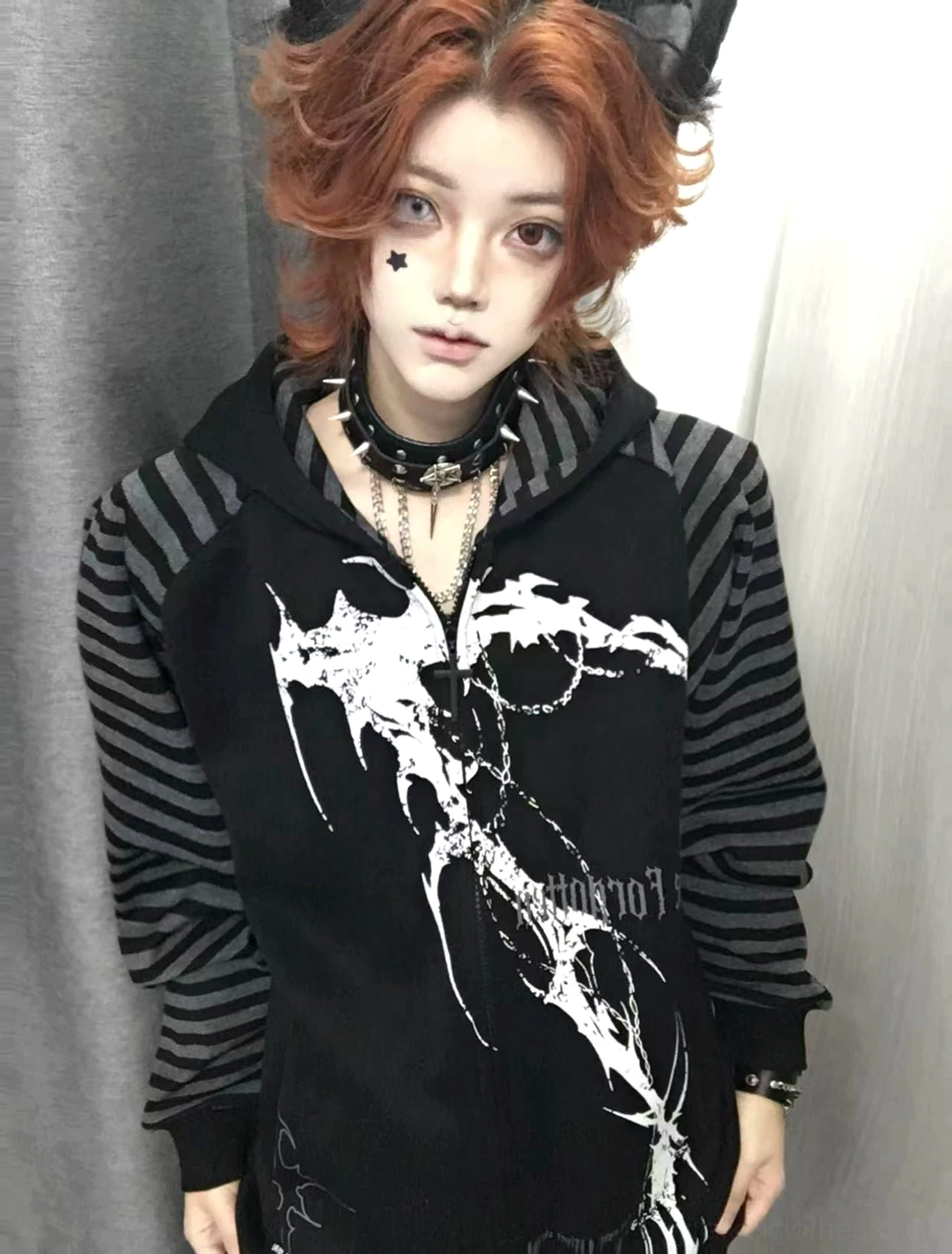 Red-haired model wearing black hooded scythe print zip coat with grey striped sleeves front half-body view plain interior backdrop Jirai Kei fashion