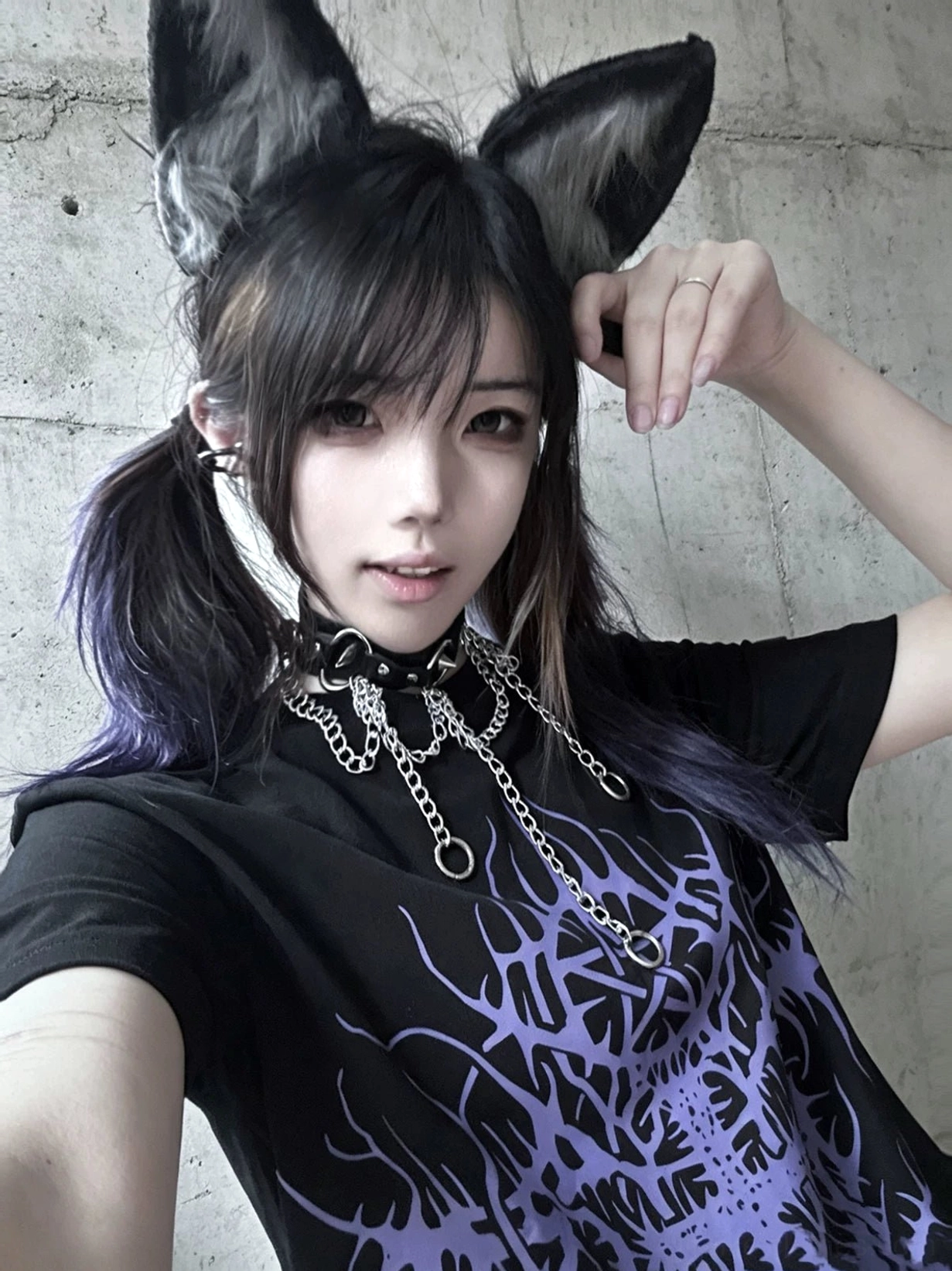 Black t-shirt with all-over purple thorn print on front, female model wearing fox ear hairpiece and chain choker, selfie view from concrete wall interior