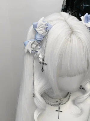 Blue-white winged bat bow hair clip with silver cross chain pendant worn on white curly wig mannequin - front three-quarter view showing Jirai Kei subculture design